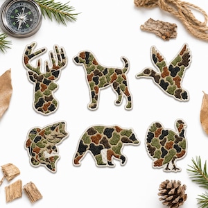 May include: A set of six embroidered fabric patches featuring wildlife silhouettes including a deer head, a dog, a flying duck, a fish, a bear, and a turkey. Each patch is filled with a multi-colored camouflage pattern in shades of green, brown, and black.