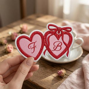 May include: A heart-shaped embroidered patch featuring two connected pink hearts with red borders. The left heart displays the letter F in cursive script, while the right heart features the letter B and a decorative red ribbon bow at the top.