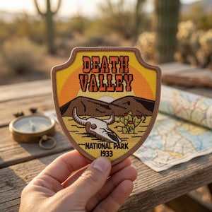 May include: A shield-shaped embroidered patch featuring a desert landscape with a skull and cactus. The text reads DEATH VALLEY NATIONAL PARK 1933. The design includes a yellow sun, brown mountains, and a tan border on a fabric backing.