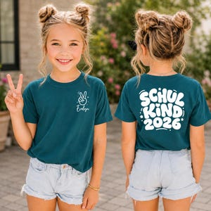 Peut inclure: Un t-shirt bleu sarcelle foncé avec un motif blanc sur la poitrine gauche représentant un signe de paix et le prénom Evelyn. Le dos du t-shirt affiche le texte blanc Schul Kind 2026 dans une police arrondie avec des petites étoiles.