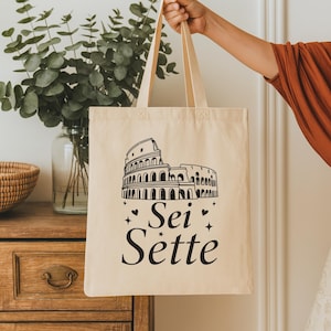 May include: Beige tote bag featuring a black illustration of the Colosseum and the text "Sei Sette." The bag has long handles and is held up by a person. The background includes a wooden cabinet and greenery in a glass vase.