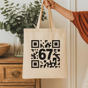 May include: A beige tote bag with a large black QR code and the number "67" printed on the front. The bag is held up by its handles, with a neutral background.