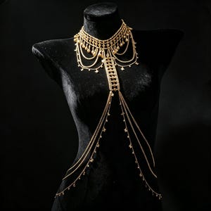 May include: A gold-toned body chain featuring a choker-style necklace with dangling teardrop charms, connected to a central vertical chain panel that extends downward into multiple draped chains adorned with small star-shaped metal accents.