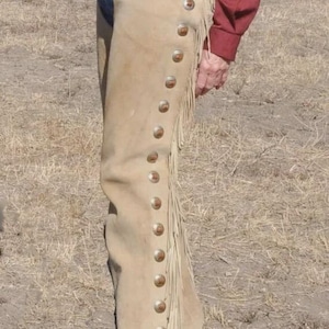 Puede incluir: Chaps de ante color beige con botones metálicos decorativos y flecos. Los chaps se llevan con un cinturón de cuero marrón y una camisa roja de manga larga. Ropa de estilo western.
