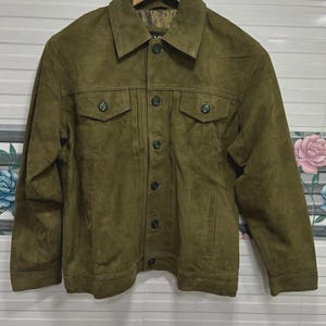 May include: Olive green suede jacket with a button-up front, two chest pockets, and two side pockets. The jacket has a classic collar and button details on the pockets. The jacket is hanging on a wooden hanger.