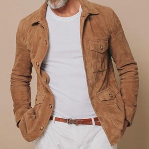 May include: A tan suede jacket with four front pockets and button closures. The jacket is worn over a white t-shirt and paired with white pants and a brown leather belt. The jacket has a classic, tailored design.