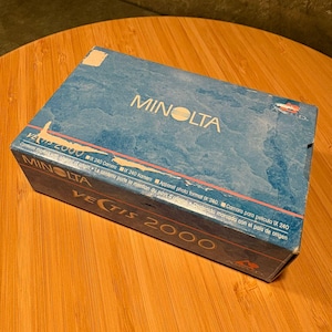 Könnte beinhalten: Blaue Minolta Vectis 2000 Kamerabox mit weißer Schrift. Die Box enthält Text in mehreren Sprachen. Die Box steht auf einer Holzoberfläche. Es ist ein Vintage-Artikel.