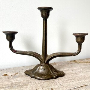 May include: A three-armed metal candelabra with a dark, weathered bronze finish. The piece features a central stem and two symmetrical side arms, each ending in a circular candle cup. The base is wide and flared with a visible stamped mark.