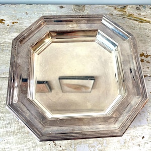 Op de afbeelding: Een achthoekig dienblad van zilverkleurig metaal met een glad, reflecterend midden en een brede, getrapte rand. Het dienblad heeft een gepolijste afwerking met zichtbare sporen van aanslag en slijtage langs de randen, wat zorgt voor een metaalachtige glans.