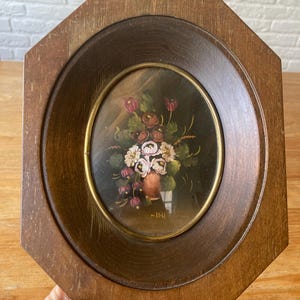 Puede incluir: Pintura floral enmarcada ovalada. La pintura presenta un ramo de flores blancas, rosas y rojas en un jarrón marrón. La pintura está sobre un fondo oscuro y enmarcada por un óvalo dorado y un marco octogonal de madera.