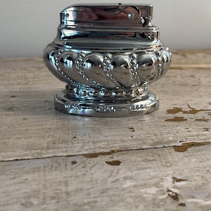 May include: A chrome lighter with a decorative, rounded base and a hinged top. The base has a repeating pattern of rounded shapes and beaded accents. The lighter is on a distressed wooden surface.