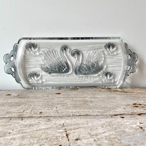 Pode incluir: Uma bandeja de servir retangular de vidro transparente com um design moldado de dois cisnes voltados um para o outro no centro, rodeados por quatro motivos de nenúfares nos cantos, com alças decorativas recortadas em ambas as extremidades.