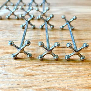 May include: A collection of silver-toned metal chopstick rests, each featuring a long, slender central rod connected to two X-shaped ends. Each end is composed of four small spheres arranged in a cross pattern, providing a stable base.