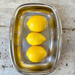 Peut inclure: Un plateau rectangulaire en acier inoxydable aux coins arrondis contenant trois citrons jaune vif disposés en ligne verticale. La surface métallique du plateau est réfléchissante et lisse, contrastant avec la peau texturée et poreuse des citrons.