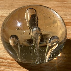 Könnte beinhalten: Ein klarer, runder Glasbriefbeschwerer mit drei tropfenförmigen Einschlüssen. Der Briefbeschwerer hat einen leichten Bernsteinton und liegt auf einer Holzoberfläche. Die Einschlüsse sind transparent.