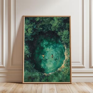 Op de afbeelding: Ingelijste kunstdruk met een cenote, een natuurlijke zinkgat, met twee figuren die in turquoise water zwemmen. Weelderig groen gebladerte omringt het water en creëert een levendige, natuurlijke scène. Het kunstwerk wordt gepresenteerd in een houten lijst.