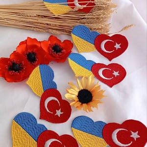 Può includere: Toppe ricamate a forma di cuore con le bandiere di Ucraina e Turchia. I cuori ucraini sono blu e gialli, mentre i cuori turchi sono rossi con una mezzaluna e una stella bianche. Papaveri rossi, un girasole e grano.