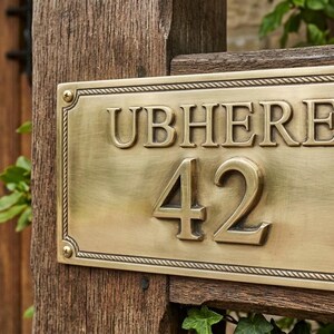 Peut inclure: Une plaque de maison rectangulaire en laiton avec le texte UBHERE et le numéro 42 en relief. La plaque présente une bordure décorative en forme de corde et est fixée à chaque coin par des vis métalliques visibles sur un poteau en bois.
