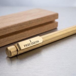 Op de afbeelding: Een goudkleurige metalen pen met een getextureerde, kruislings gearceerde houder en een platte clip gegraveerd met de tekst 2 PRECISION. De pen ligt naast een rechthoekige houten kist met een gladde afwerking en een horizontale groef.