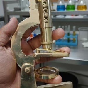 Puede incluir: Un microscopio de latón con la inscripción "Dr. DeLuca". La imagen muestra el instrumento sostenido por una mano, con un laboratorio en el fondo, incluyendo vasos de precipitados y tubos de ensayo.