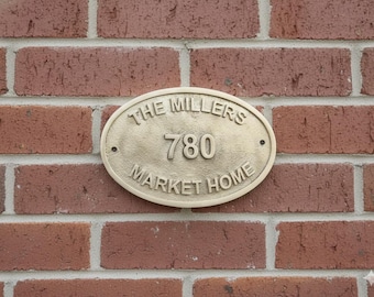 May include: An oval, gold-colored address plaque with the text "THE MILLERS 780 MARKET HOME" in a serif font. The plaque is mounted on a red brick wall.