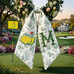 May include: A golf-themed bow with a green and white toile pattern. The bow displays the Masters logo and a large green "M". The background shows a golf course with a flag and a golf cart.