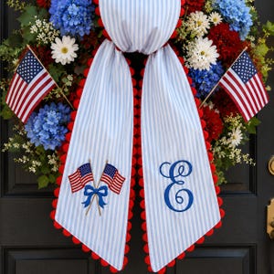 May include: A patriotic wreath with red, white, and blue flowers and greenery. The wreath features a striped fabric with red trim and an embroidered letter "E". Two small American flags are placed on either side.