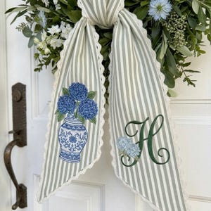 Puede incluir: Una cinta de tela a rayas verde salvia y blanco con bordes de encaje festoneado. Un lado presenta un ramo de hortensias azules bordado en un jarrón blanco y azul. El otro lado muestra una letra H grande bordada en verde oscuro con una pequeña hortensia.