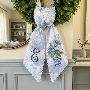 Pode incluir: Uma fita de tecido branco com padrão toile azul, apresentando uma borda de renda branca recortada. Um lado exibe a letra E bordada em azul escuro, enquanto o outro lado mostra uma ilustração impressa de hortênsias azuis e brancas num vaso.