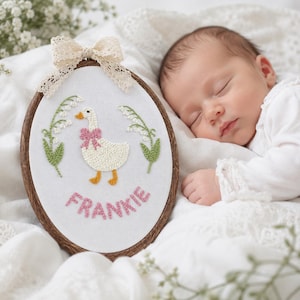 Könnte beinhalten: Ein ovaler, hölzerner Stickrahmen mit einer gestickten Gans mit rosa Schleife und dem Namen "FRANKIE". Grüne und weiße Blumenmotive umgeben die Gans. Oben befindet sich eine cremefarbene Spitzenschleife. Der Rahmen liegt neben einem schlafenden Baby.
