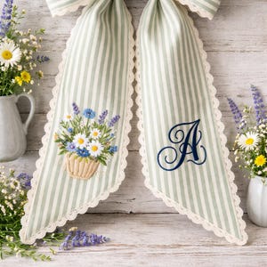 Puede incluir: Un lazo de tela a rayas verde salvia y blanco con un borde de encaje color crema. Un extremo muestra una cesta bordada de margaritas y lavanda, mientras que el otro extremo presenta una letra A grande bordada en azul oscuro en cursiva.