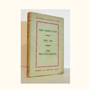 Pode incluir: Uma capa de livro vintage com uma borda vermelha e texto verde. O título é "The Simpleton, The Six, The Millionairess" de Bernard Shaw.