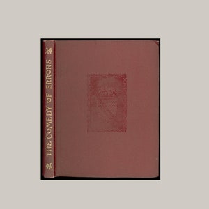 May include: A burgundy-colored book with gold lettering on the spine that reads "THE COMEDY OF ERRORS". The front cover has a decorative design in the center.