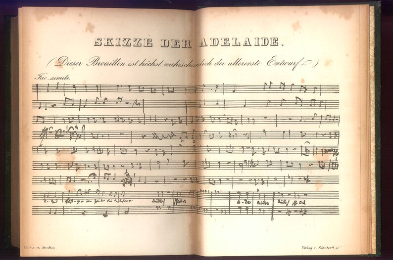 May include: Open book with aged, yellowed pages featuring handwritten musical notation. The title "SKIZZE DER ADELAIDE" is visible at the top. The text is in German. The book's spine is dark green.
