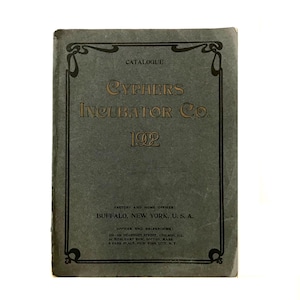 May include: A vintage catalog cover for Cyphers Incubator Co. from 1902. The cover is a muted gray-green with gold lettering and decorative borders. The text includes the company name, the year, and the factory and office locations in Buffalo, New York, and other cities.