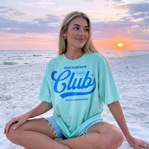 May include: A pale green t-shirt featuring the blue text "THE MOCKUP Clubs". The words "YOUR DESIGN HERE" are below the word "Clubs". The image is set on a beach with a sunset in the background.