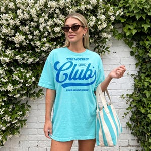 Puede incluir: Una camiseta azul claro con el texto "THE MOCKUP CLUB" en azul oscuro. La camiseta se combina con un bolso tote a rayas blancas y azul claro. El fondo presenta una pared de ladrillos blancos y vegetación.