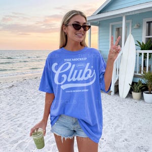 May include: A periwinkle blue oversized t-shirt featuring the white text The Mockup Club Your Design Here in a script font. The shirt has short sleeves and a crew neckline, worn with light wash denim shorts with a frayed hemline.