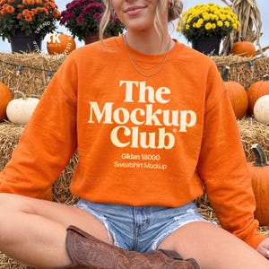 Può includere: Una felpa girocollo arancione con la scritta color crema The Mockup Club, con un testo più piccolo sottostante che recita Gildan 18000 Sweatshirt Mockup. Il capo presenta maniche lunghe e un colletto a coste, indossato con pantaloncini di jeans.