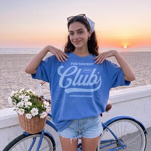 May include: A blue t-shirt with the text "The Mockup Club" and "Your Design Here" in a light blue font. The t-shirt is worn with denim shorts. A blue bicycle with a wicker basket filled with white flowers is in the foreground.