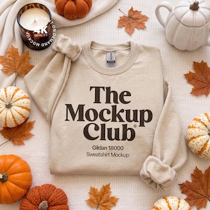 May include: A beige crewneck sweatshirt featuring the dark brown text The Mockup Club and Gildan 18000 Sweatshirt Mockup printed on the front. The garment has long sleeves with ribbed cuffs and a ribbed neckline, displayed on a textured fabric surface.