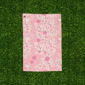 May include: Pink golf towel with a floral pattern. The towel features a repeating design of white, pink, and light blue flowers on a pink background. A metal grommet is visible at the top corner.