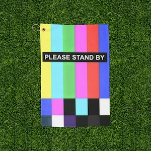 May include: A rectangular golf towel with a colorful test pattern design, featuring vertical stripes of yellow, blue, green, pink, and red. The towel has the text "PLEASE STAND BY" in a black rectangle and a metal grommet for hanging.
