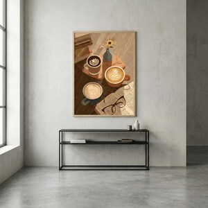 May include: A framed art print depicts three coffee cups with latte art, a book, and eyeglasses on a wooden surface. A small vase with a sunflower sits nearby. The artwork has a warm, inviting color palette.