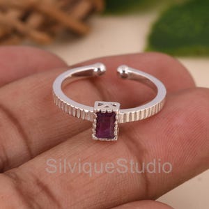 Puede incluir: Un anillo abierto de plata con una piedra preciosa rectangular de color rosa intenso engastada en una banda texturizada. La banda tiene ranuras verticales en los lados y un pequeño marco metálico decorativo alrededor de la piedra central. El texto SilviqueStudio es visible.