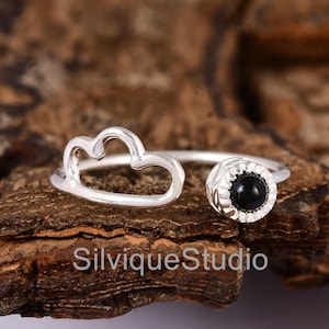 Peut inclure: Une bague argentée avec un motif en forme de nuage et une pierre noire. La bague a un design ouvert, avec la forme de nuage d'un côté et la pierre précieuse de l'autre. La bague est posée sur un fond marron. Le texte "SilviqueStudio" est visible.