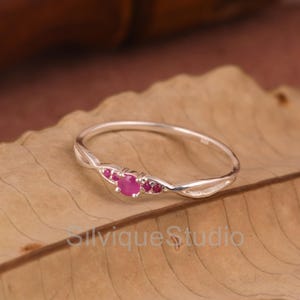 Puede incluir: Un delicado anillo de plata con una gema central de rubí redondo, flanqueada por pequeños acentos de rubí. La banda tiene un diseño retorcido, añadiendo un toque de elegancia. El anillo se muestra sobre una hoja marrón.