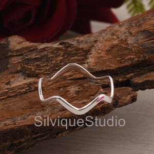 May include: A silver ring with a wave-like design, displayed on a piece of dark wood. The ring has a smooth, polished finish and a distinctive, flowing shape. The text "SilviqueStudio" is visible on the wood.