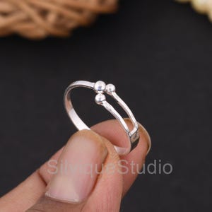 May include: A silver ring with a unique design. The ring features a split band with three small, round accents. The ring is held by a hand against a dark background. The text "Silvique Studio" is visible.
