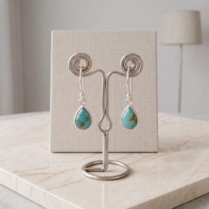 Puede incluir: Un par de pendientes colgantes de plata con piedras de turquesa en forma de lágrima y vetas marrones. Los pendientes cuelgan de ganchos de plata y se exhiben en un soporte de metal plateado con dos espirales en la parte superior.